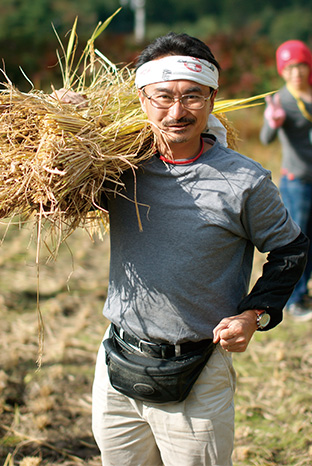 刈り取った稲を担ぐ、代表理事の曽根原久司さん NPO法人「えがおつなげて」│山梨県北杜(ほくと)市 取材/原口真吾(本誌) 写真/「えがおつなげて」提供