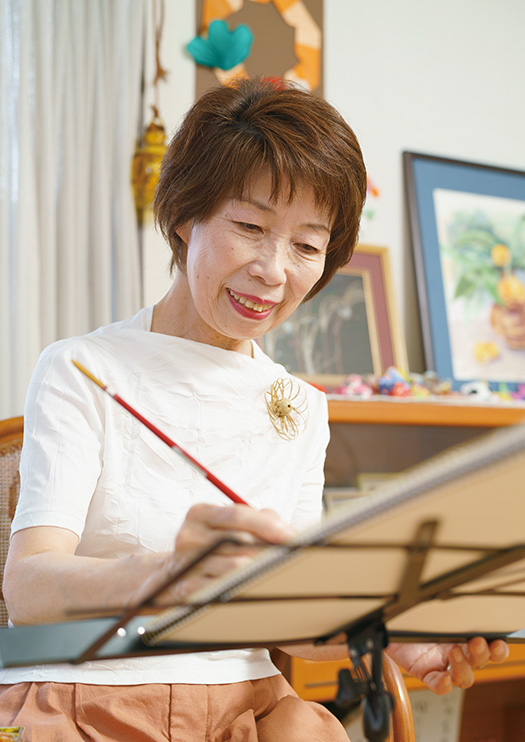 三井眞知子(みつい・まちこ)さん│71歳│広島県竹原市 自宅のアトリエで絵を描く三井さん。後ろの壁や棚には、これまで描いた水彩画のほか、折り紙なども飾られている 取材・写真/永谷正樹
