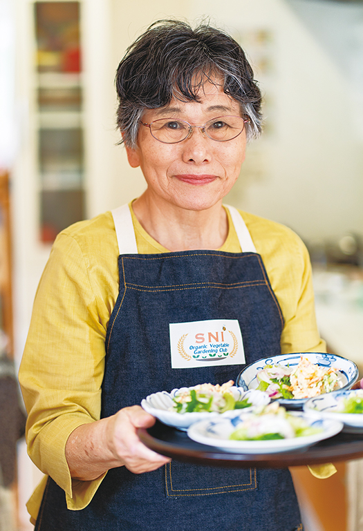 自ら作った昼食をお盆に載せて微笑む中村さん。SNオーガニック菜園部のエプロンをつけて